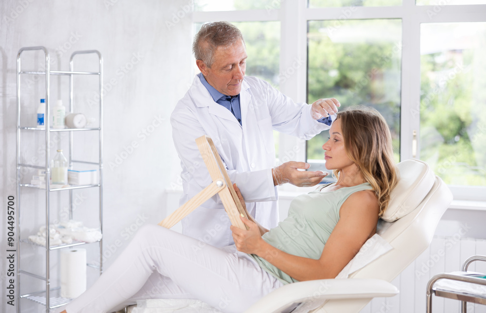 Obraz premium Female patient of cosmetology office sitting in chair with mirror in hands and discussing with mature male doctor need for rejuvenation procedure laser skin resurfacing forehead