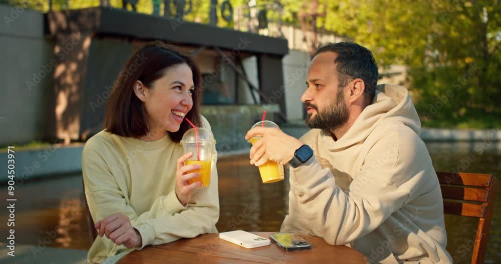 Happy brunette girl and her boyfriend with a beard are talking while sitting at a table in a cafe and drinking orange juice during their date in the park Happy brunette girl and her boyfriend with a beard are talking while sitting at a table in a cafe and drinking orange juice during their date in the park