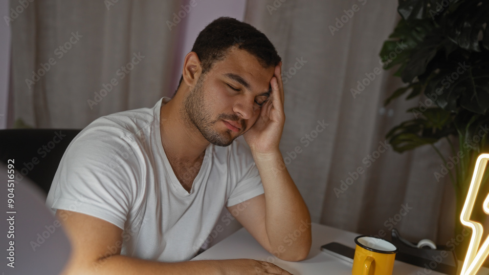 Young hispanic man sitting in a living room with a tired expression, resting his head on one hand, wearing a white shirt, with a yellow mug on the table. Young hispanic man sitting in a living room with a tired expression, resting his head on one hand, wearing a white shirt, with a yellow mug on the table.