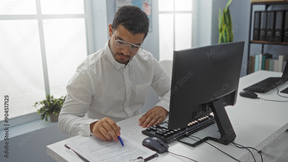 Hispanic man working at a desk in an office with a computer and documents, creating an atmosphere of productivity and diligence. Hispanic man working at a desk in an office with a computer and documents, creating an atmosphere of productivity and diligence.