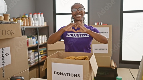 Smiling woman wearing glasses and volunteer shirt forming heart gesture in donation center with boxes and supplies in background.