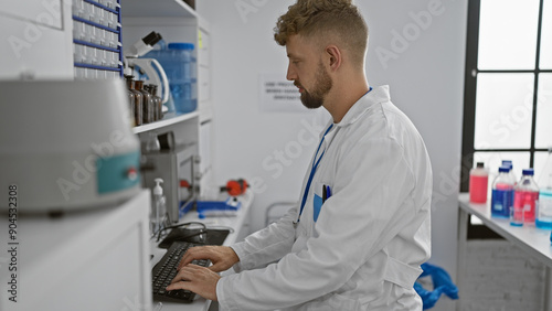 A young, caucasian male scientist with a beard works at a computer inside a modern laboratory.