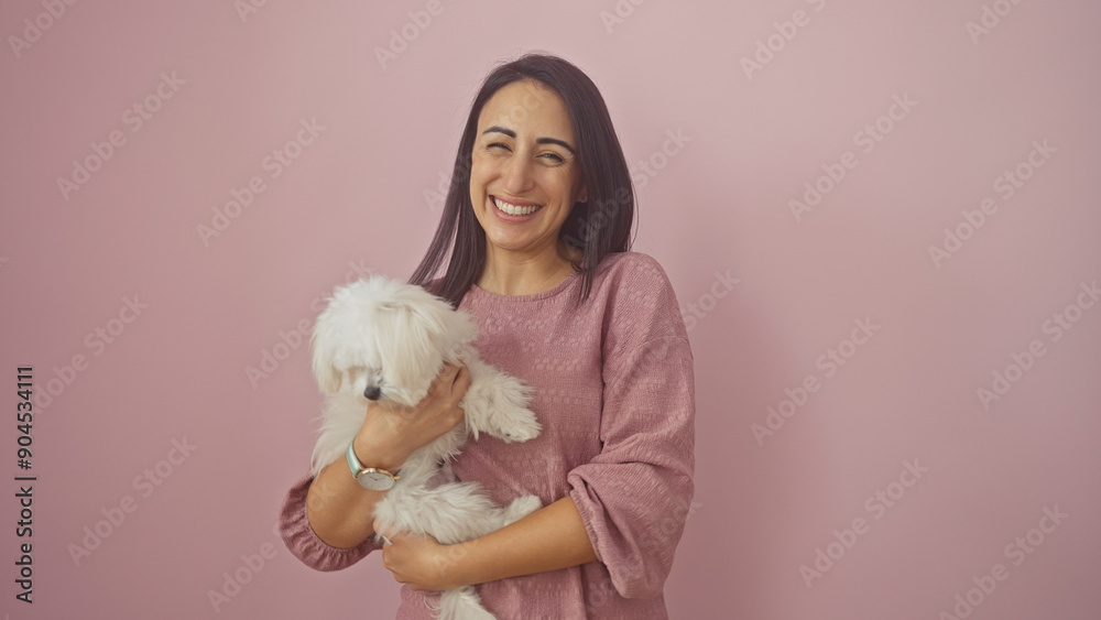 A happy hispanic adult woman in a pink blouse holding a white bichon maltese dog against a pink background