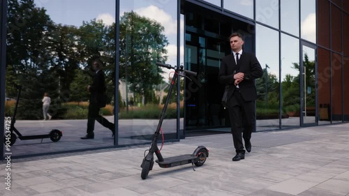 A man dressed in a black suit and tie exits a business center. The businessman steps onto an electric scooter and rides out of frame, with a modern building in the background.