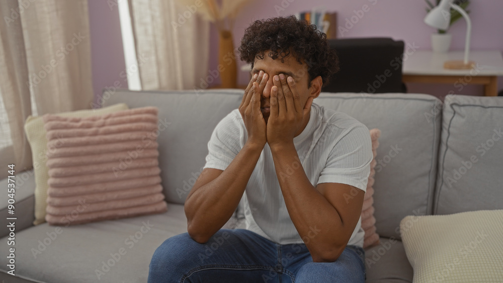 Young hispanic man sitting on a sofa in a living room with his hands covering his face, suggesting frustration or stress in a home environment Young hispanic man sitting on a sofa in a living room with his hands covering his face, suggesting frustration or stress in a home environment