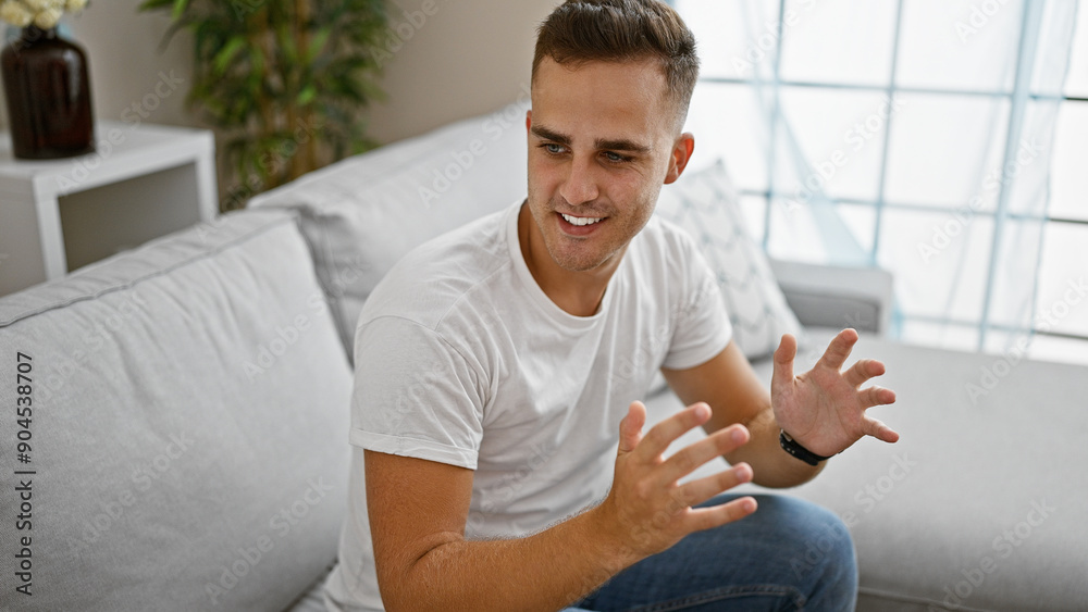 Casual young hispanic man smiling in bright living room interior wearing white shirt jeans and watch Casual young hispanic man smiling in bright living room interior wearing white shirt jeans and watch