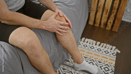 Young caucasian man in shorts sitting indoors holding his knee as if experiencing pain or discomfort.