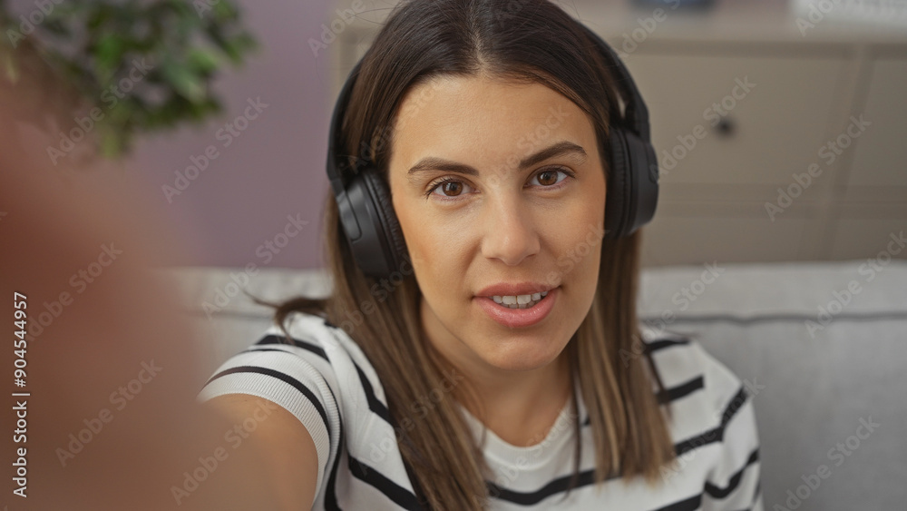 A young brunette woman wearing headphones takes a selfie in a modern living room with a cozy atmosphere. A young brunette woman wearing headphones takes a selfie in a modern living room with a cozy atmosphere.