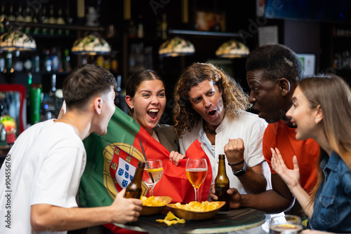 Group of cheerful emotional people of different ages and nationalities cheering for favorite sports team in pub, waving national flag of Portugal while watching football match on TV..