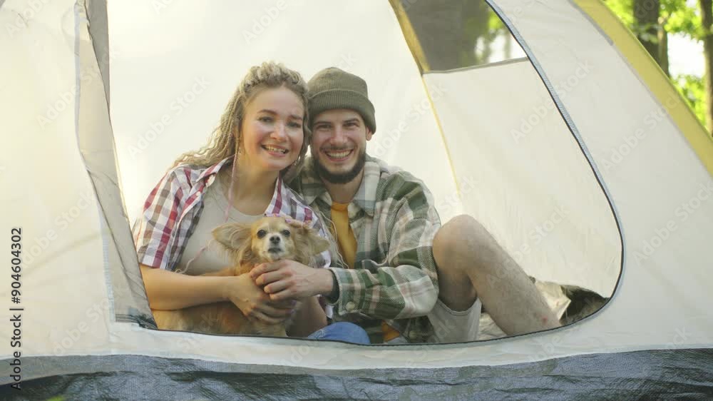 A couple in love enjoys relaxing in a tent in the forest while their playful chihuahua dog rests at their feet, looking at the camera and smiling