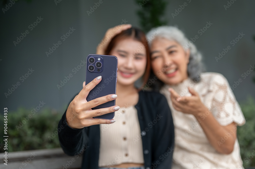 Fototapeta premium A lovely Asian granddaughter and her grandma are sitting on a bench and taking a selfie together.