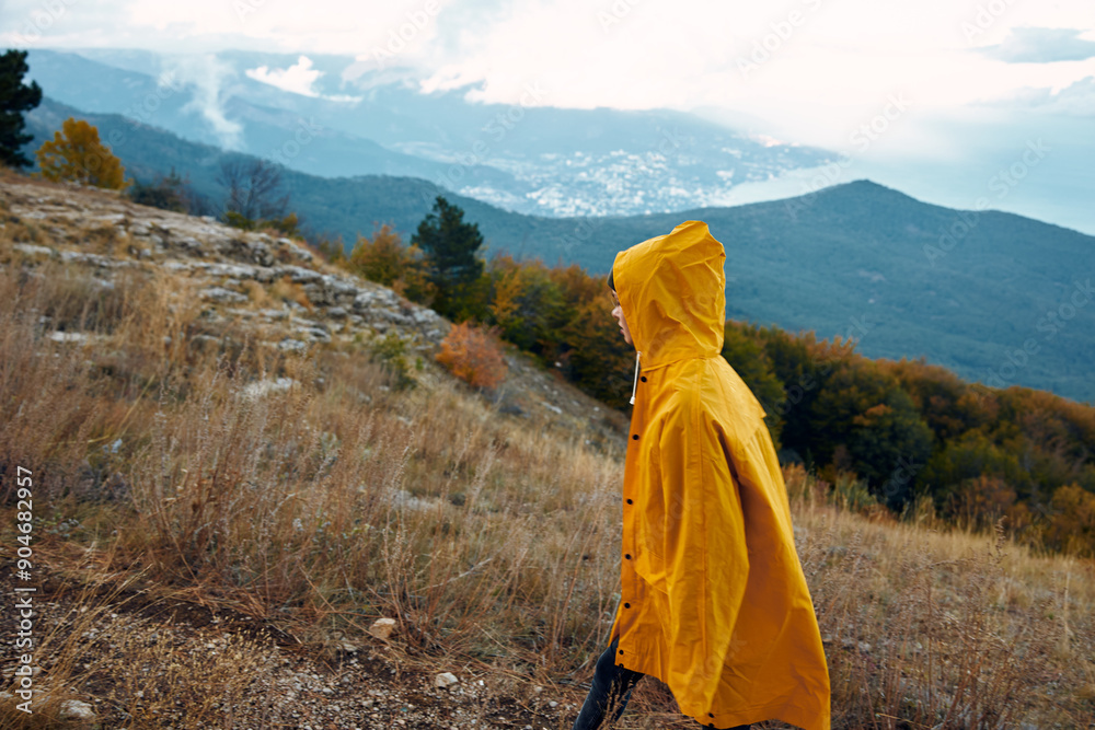 A person in a yellow raincoat standing on a hillside admiring the majestic mountain landscape in the background A person in a yellow raincoat standing on a hillside admiring the majestic mountain landscape in the background