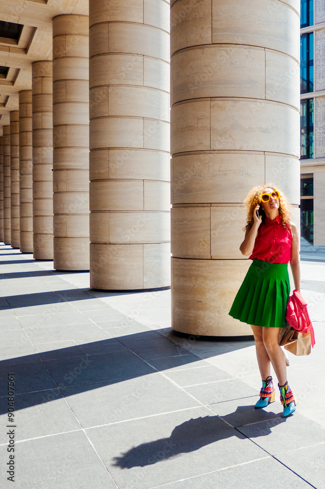 Content fashion female with bag walking on pavement near stone columns in sunlight and talk cell phone. the concept of freedom of tourism and fashion business