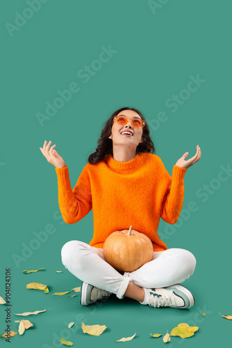 Young Asian woman in fall clothes with pumpkin and leaves sitting on green background