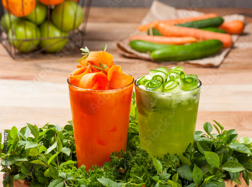 Beautifully decorated freshly prepared carrot and cucumber juice glasses on a tray filled with various edible greens on wooden table  with other fruits and vegetables in the background