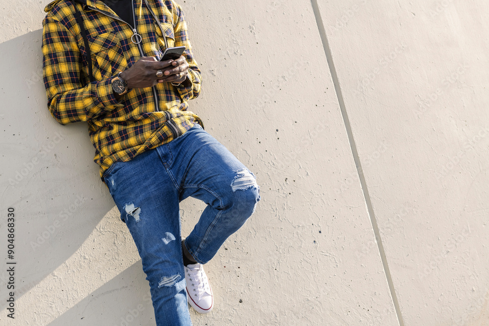 unrecognizable young african man writing a message on his phone leaning on a modern concrete structure, concept of lifestyle and technology unrecognizable young african man writing a message on his phone leaning on a modern concrete structure, concept of lifestyle and technology