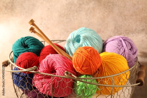 Many soft skeins of yarn and knitting needles in metal basket on light brown background, closeup photo