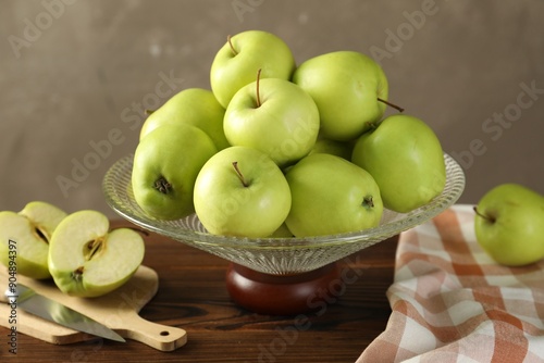 Glass vase with fresh green apples, cutting board and knife on wooden table against gray background, closeup