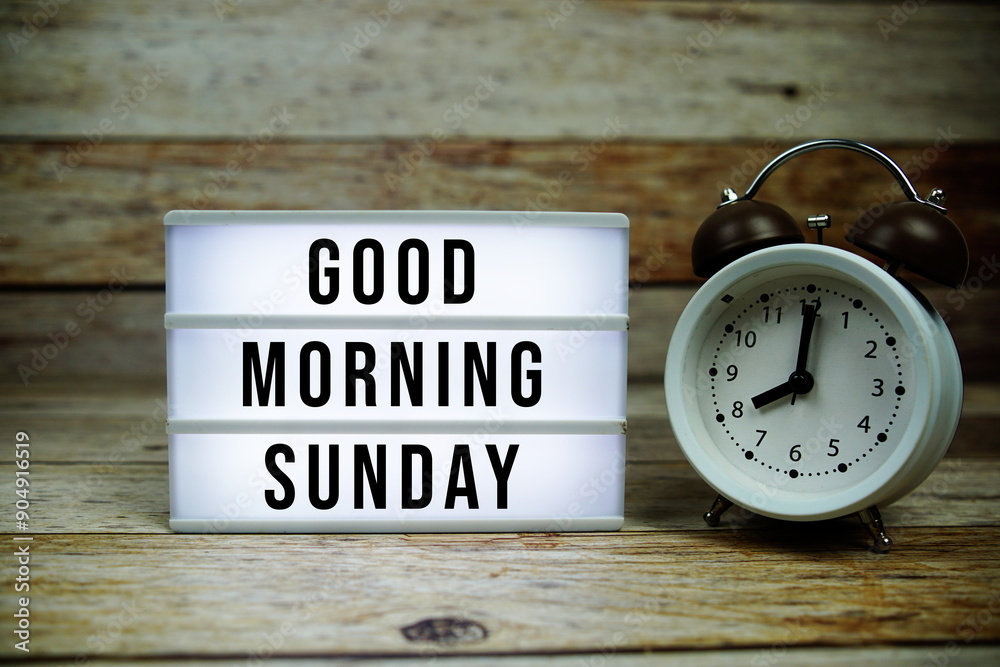 Good Morning Sunday text on LED lightbox with alarm clock on wooden background