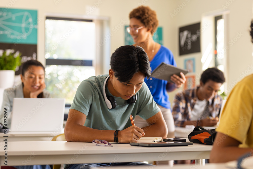 In high school, teenage boy writing in notebook while teacher with tablet observes In high school, teenage boy writing in notebook while teacher with tablet observes