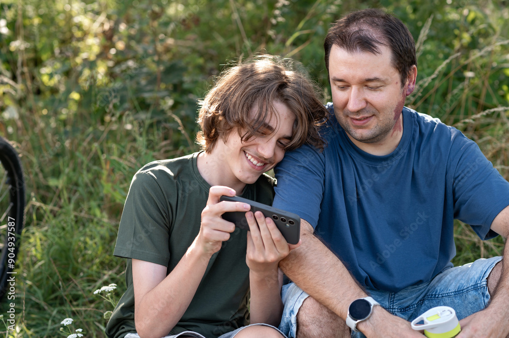 Teenager boy and his father sitting in nature and looking at smartphone screen, talking and smiling Teenager boy and his father sitting in nature and looking at smartphone screen, talking and smiling