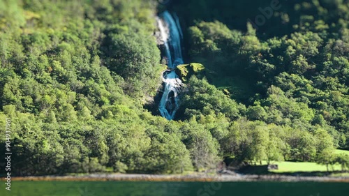 A tilt-shift video captures a serene waterfall on the coast of the Naeroy fjord forest, surrounded by vibrant greenery. The blurred edges create a toy-like appearance, enhancing the natural beauty. photo