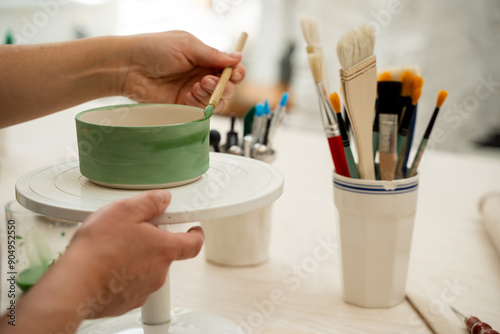 Fototapeta Naklejka Na Ścianę i Meble -  Girl is decorating a clay product with bright green color. Using a brush to paint a ceramics bowl. Learning how to paint a handmade pottery project.