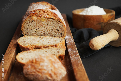Fresh sliced ​​bread on a wooden tray next to a bowl of flour and a wooden rolling pin on a dark background