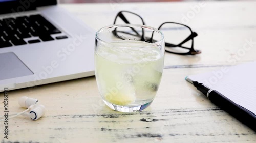 A hand drops an effervescent tablet into a glass of water on a wooden desk surrounded by office supplies, suggesting a home workspace or study area. The scene implies that the user is managing fatigue photo