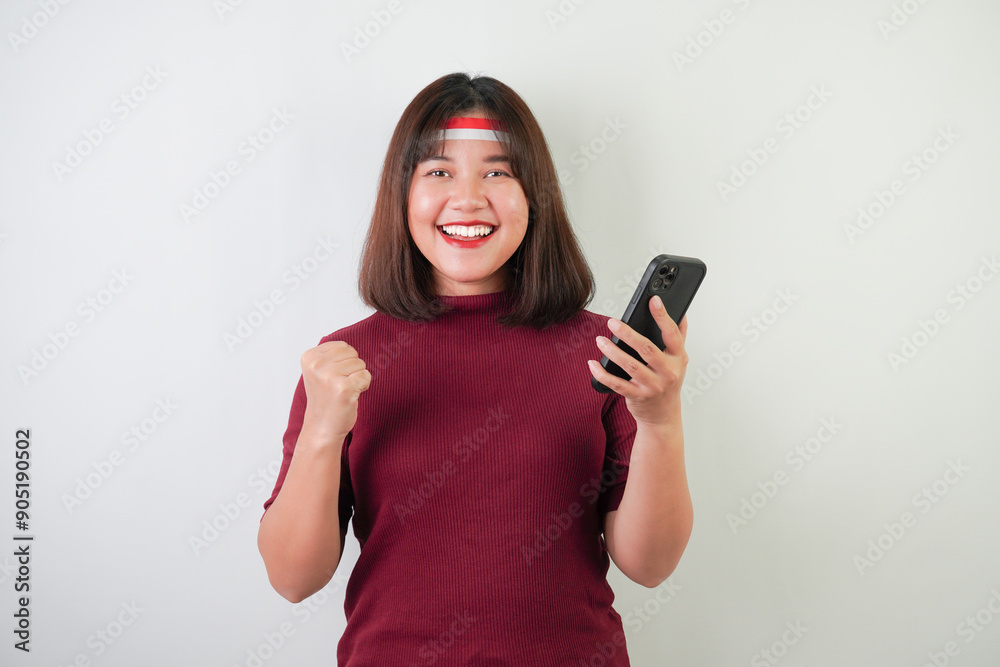 Excited young asian woman wearing Indonesian Flag headband is watching vertical smartphone screen for advertising, clenched fist gesture, isolated over white background. Indonesian Independence Day. Excited young asian woman wearing Indonesian Flag headband is watching vertical smartphone screen for advertising, clenched fist gesture, isolated over white background. Indonesian Independence Day.
