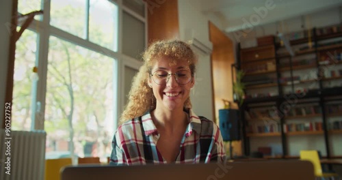 Happy girl with curly hair wearing round glasses sits in front of a laptop and waves her hand while communicating via video conference in the library