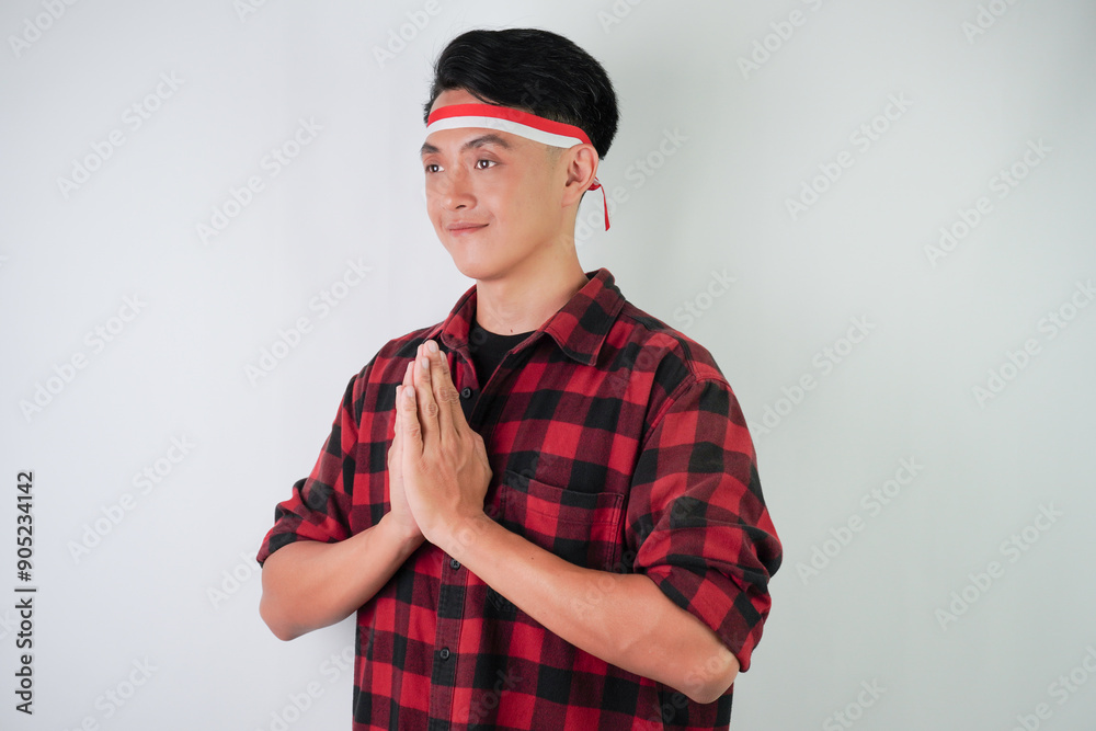 Fototapeta premium Excited young asian man wearing Indonesian flag headband, smiling expression with hands pressed together gesture, isolated over white background. Concept for Indonesian Independence Day.