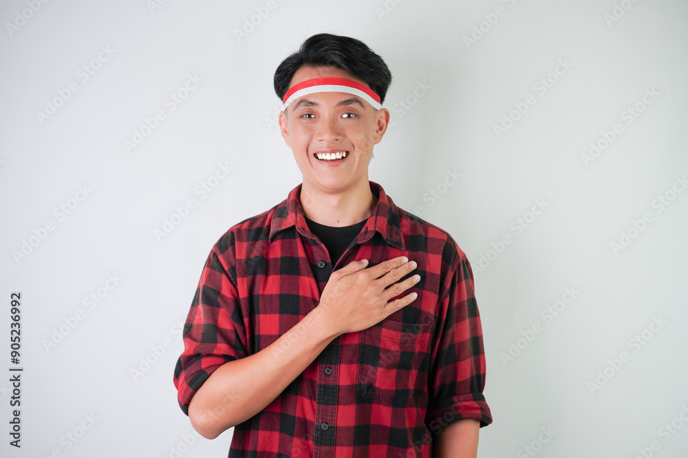 Fototapeta premium Excited young asian man wearing Indonesian flag headband, smiling expression with hand palm on heart saluting gesture, isolated over white background. Concept for Indonesian Independence Day.