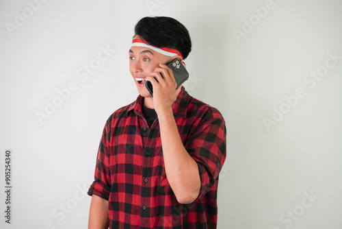 Excited young asian man wearing Indonesian Flag headband is talking using smartphone, receiving call from someone, isolated over white background. Indonesian Independence Day.