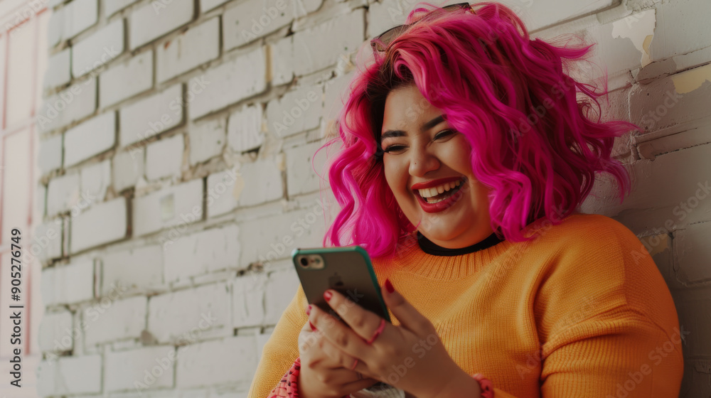 Fototapeta premium A person with vibrant pink hair dressed in a yellow sweater laughs while looking at their phone, leaning against a textured brick wall.