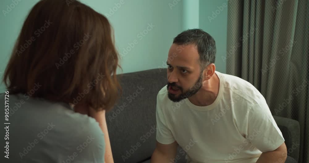 Over the shoulder Furious brunette man with a beard in a white T-shirt shouts at his girlfriend while swearing on the couch at home Over the shoulder Furious brunette man with a beard in a white T-shirt shouts at his girlfriend while swearing on the couch at home