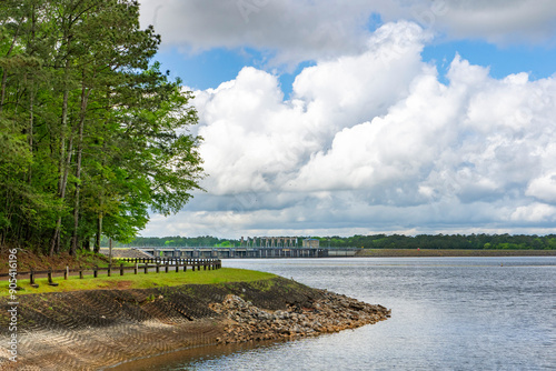 A wide-angle view of the West Point Lake Dame from Eagleview Park in Georgia against a backdrop of blue sky with large white clouds. Birds can be seen soaring in the sky.