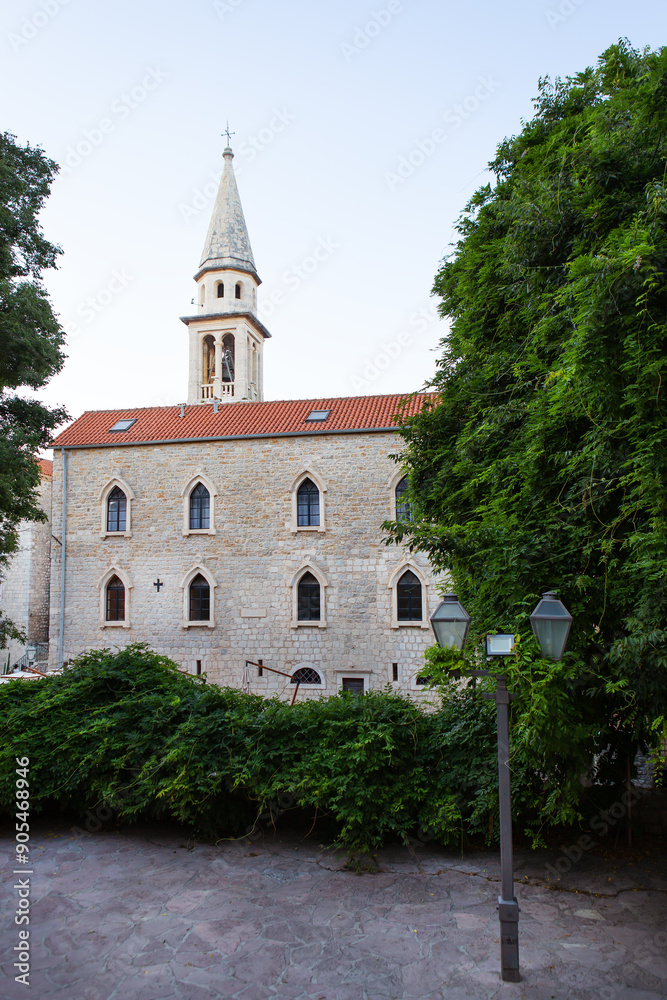 Naklejka premium An old stone church with a bell tower stands amidst lush green trees and a cobblestone courtyard adorned with street lamps.