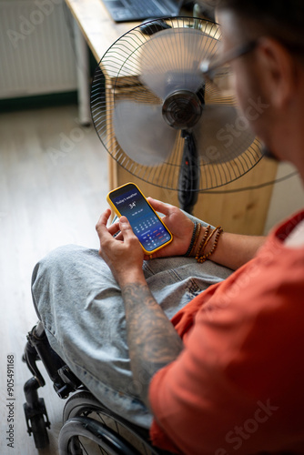 Disabled paralyzed man in wheelchair checking weather forecast in mobile app on smartphone, sits by ventilator fan at home during extreme summer heat wave, suffers from lack of air conditioning. photo