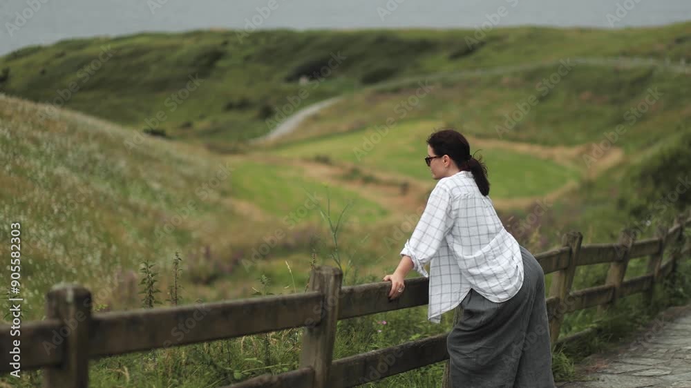 Attractive girl stands by a wooden fence on a hiking trail, enjoying the beautiful view of the northern landscapes of the ocean coast and green meadows, enjoying nature and freedom, eco tourism, wild Attractive girl stands by a wooden fence on a hiking trail, enjoying the beautiful view of the northern landscapes of the ocean coast and green meadows, enjoying nature and freedom, eco tourism, wild