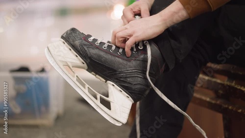 A close-up view of someone loosening the laces of a black ice skate with two hands, with a blue skating shoe visible in a transparent container in the background photo