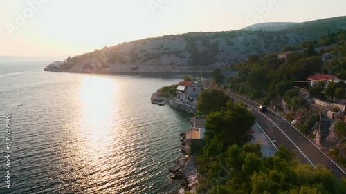 Passenger car driving alongside seashore. Sun reflects on sea bay water and road follows hill slope shape near mountains on shoreline aerial view photo