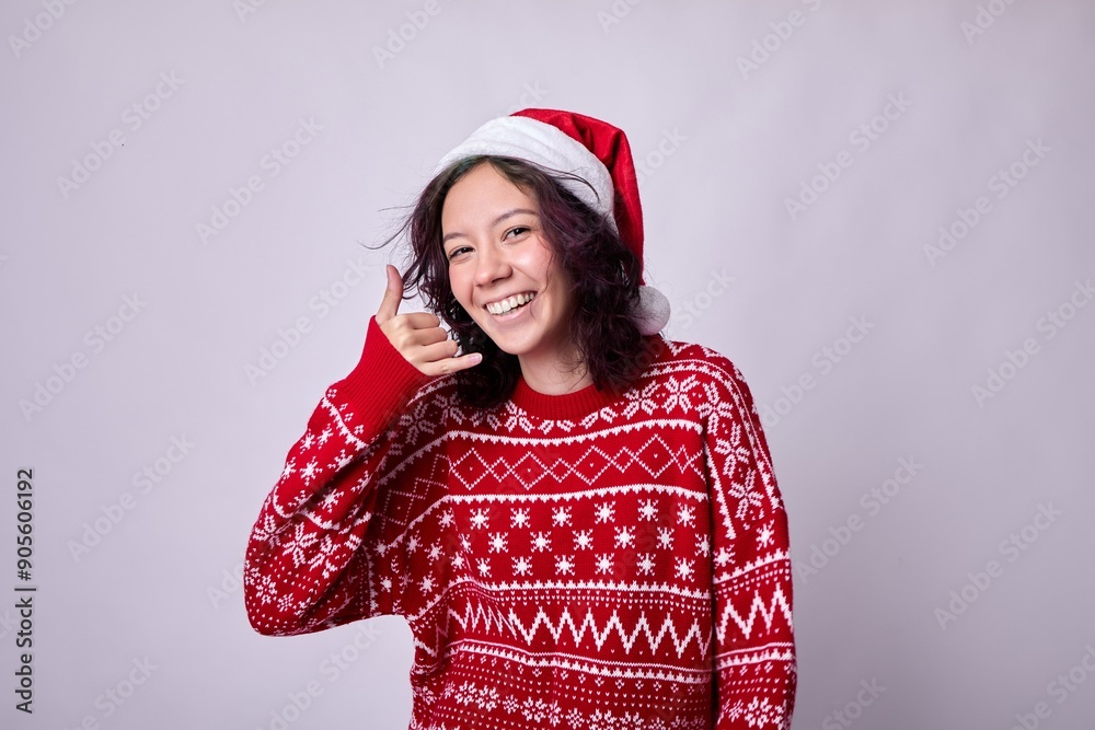 Young woman in a cozy red sweater and Santa hat smiles joyfully as she makes a playful call me gesture, exuding a festive vibe with delicate snowflakes adorning the gray background. Young woman in a cozy red sweater and Santa hat smiles joyfully as she makes a playful call me gesture, exuding a festive vibe with delicate snowflakes adorning the gray background.