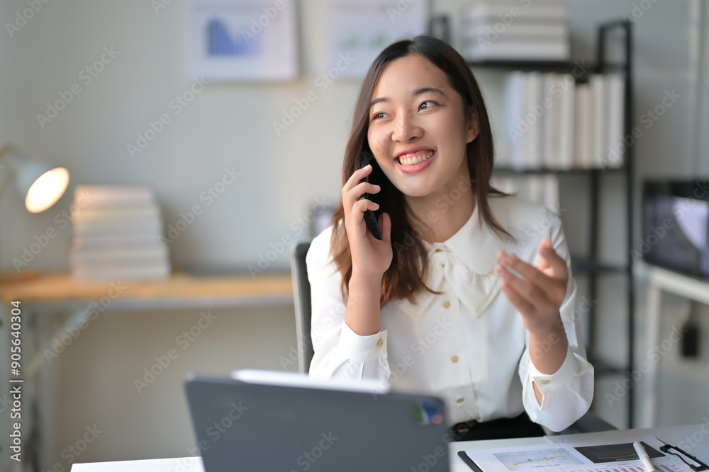 Young Professional Woman Smiling While Talking on Phone in Modern Office Setting with Laptop and Documents Young Professional Woman Smiling While Talking on Phone in Modern Office Setting with Laptop and Documents