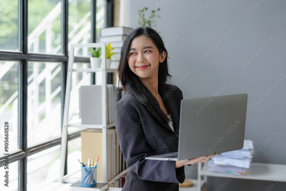 Young professional woman smiling and holding a laptop while standing in a modern office Young professional woman smiling and holding a laptop while standing in a modern office