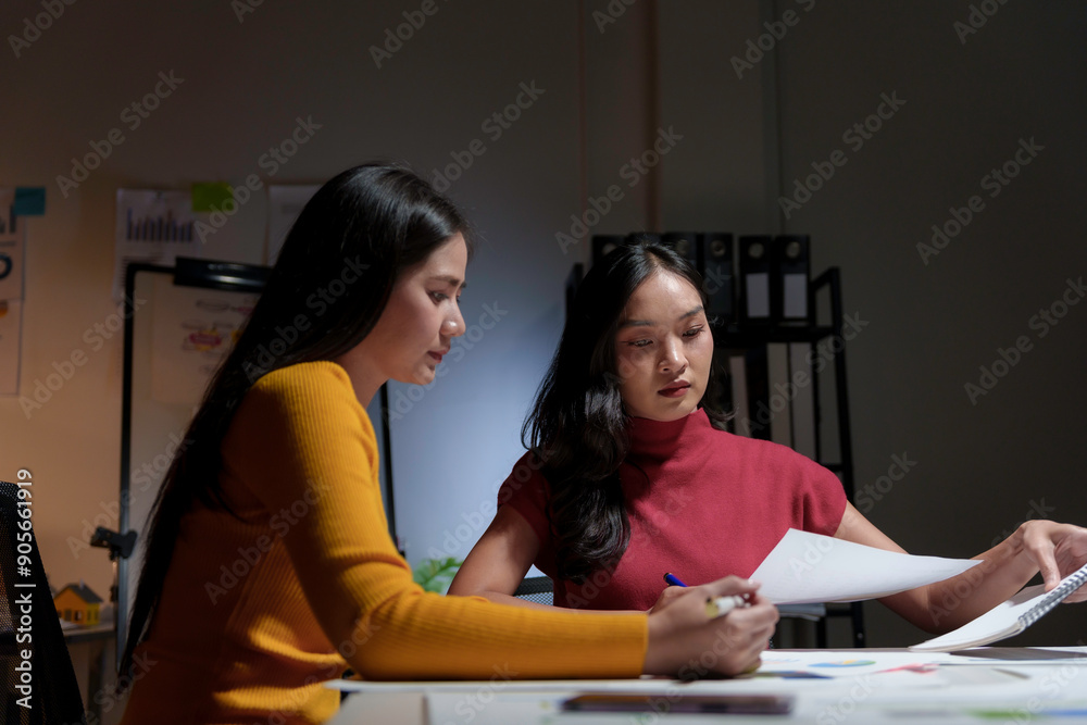 Two young businesswomen working late in the office, reviewing paperwork and analyzing financial charts Two young businesswomen working late in the office, reviewing paperwork and analyzing financial charts