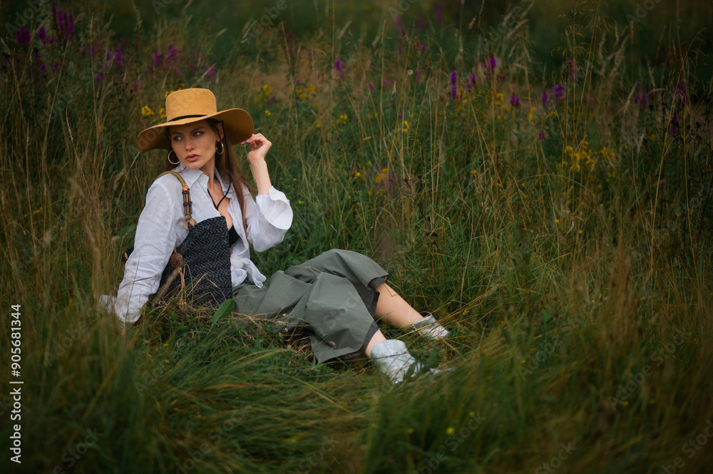 Fashionable confident woman wearing trendy summer hat, white linen shirt, green skirt, holding wicker straw bag, sitting on grass. Outdoor full-length fashion portrait. Copy, empty space for text Fashionable confident woman wearing trendy summer hat, white linen shirt, green skirt, holding wicker straw bag, sitting on grass. Outdoor full-length fashion portrait. Copy, empty space for text