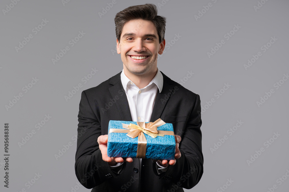 Handsome Businessman In Suit Gives Gift Box Present To Camera. Man is happy, smiling on grey office wall background. Positive mood, holiday, bonus, birthday concept. Handsome Businessman In Suit Gives Gift Box Present To Camera. Man is happy, smiling on grey office wall background. Positive mood, holiday, bonus, birthday concept.
