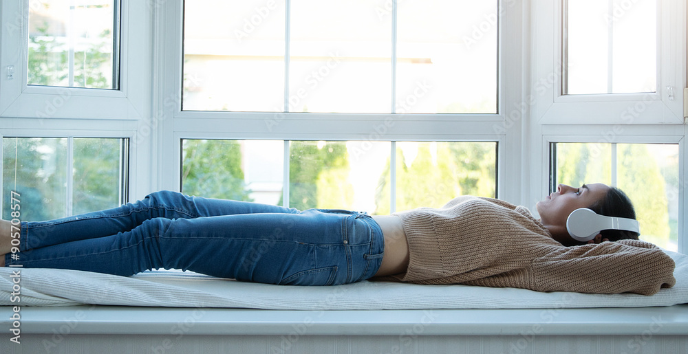 Calm confident thoughtful cute lovely young modern woman in wireless headphones laying down on big windowsill relax and dreaming about something. Girl listening music, podcast or online book Calm confident thoughtful cute lovely young modern woman in wireless headphones laying down on big windowsill relax and dreaming about something. Girl listening music, podcast or online book