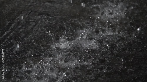 A black and white photo of rain falling on a dark surface. Scene is somber and melancholic, as the rain seems to be falling heavily and the surface appears to be desolate